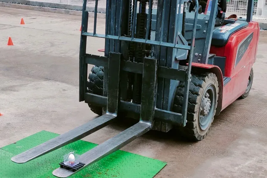 Montacargas con un huevo sobre la uña frente a una placa marcada en el piso, en una prueba de control fino de la carga