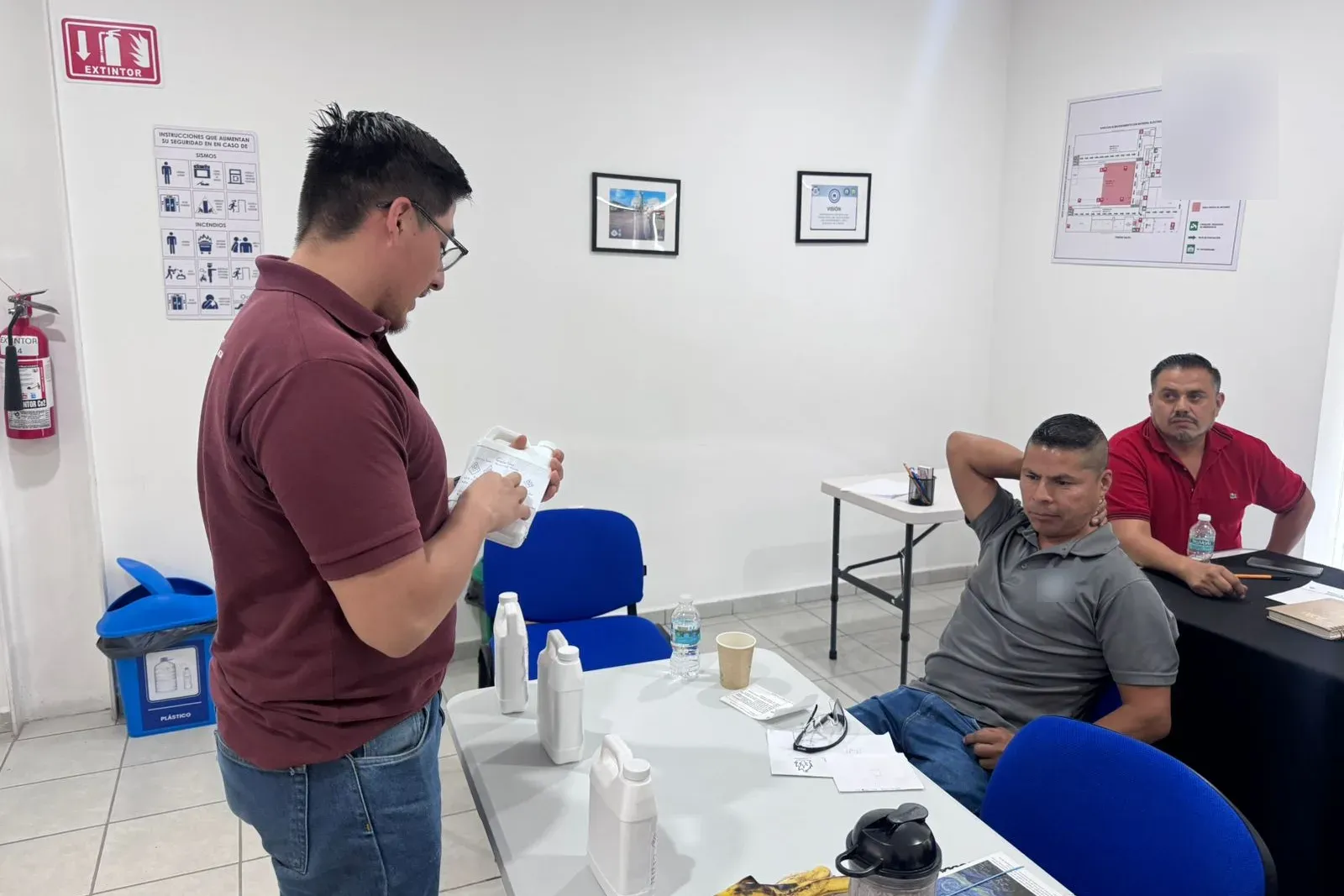 Instructor revisa la etiqueta de peligros escrita en una garrafa de prueba mientras dos participantes observan en el aula