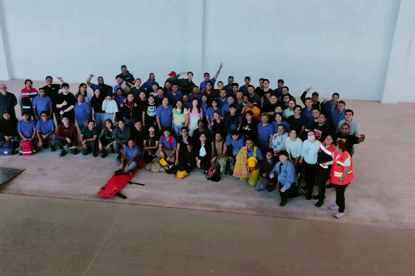 Grupo numeroso de brigadistas con camilla rígida, botiquines y equipo de bombero tras una capacitación en una nave