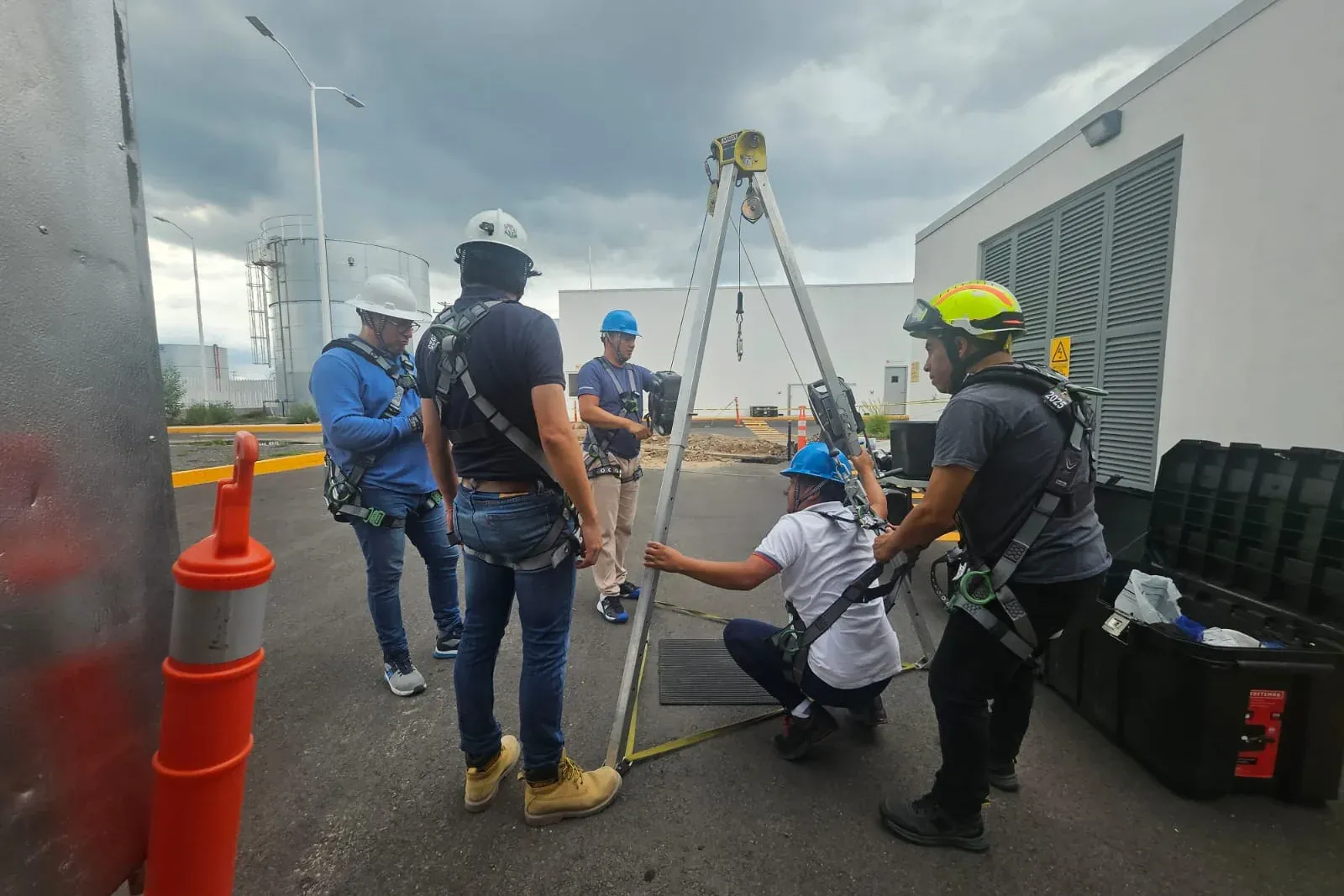 Participante agachado bajo un trípode conecta su arnés a la línea de vida mientras compañeros con casco y arnés lo asisten