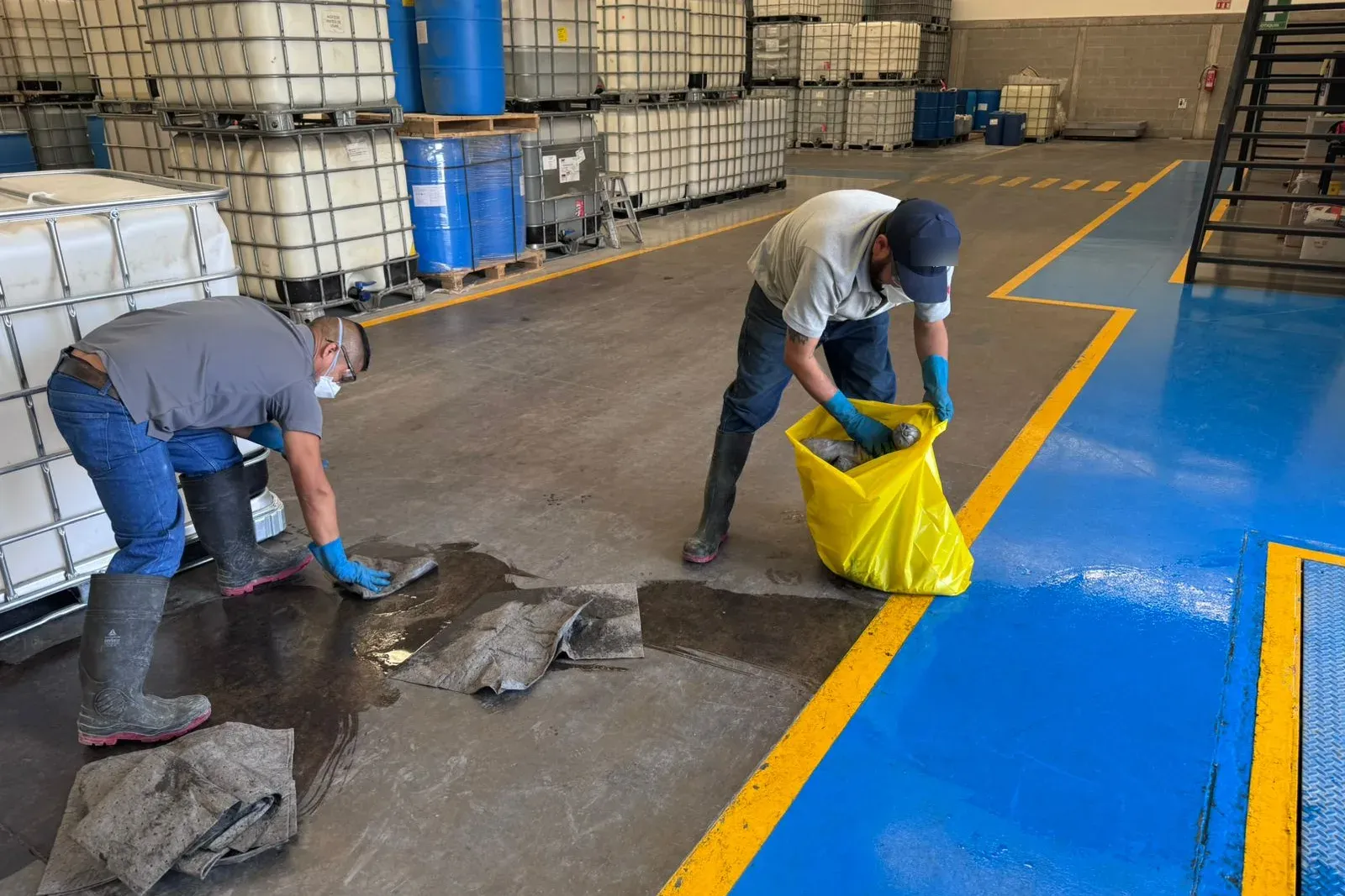 Dos trabajadores con guantes, mascarilla y botas recogen paños absorbentes empapados y los meten en una bolsa de residuos
