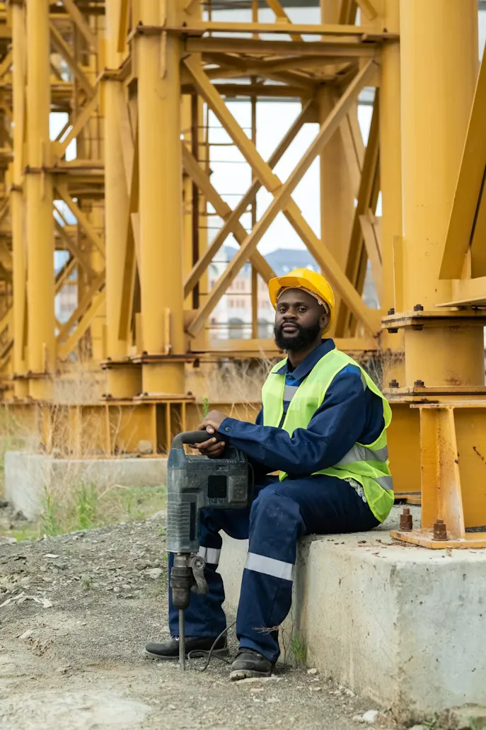 Trabajador industrial con chaleco y casco sentado junto a estructura metálica
